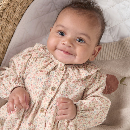 A baby girl lies in a changing basket wearing a pink dress with a ruffled collar.