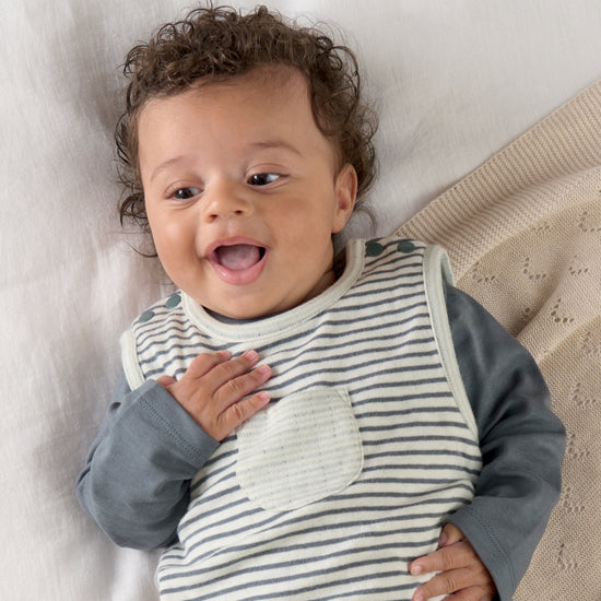 A smiling baby boy is lying down wearing striped dungarees with a blue bodysuit underneath.