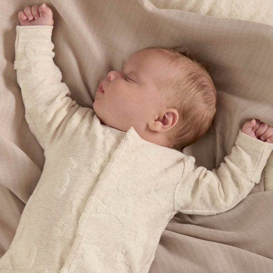 A baby is lay sleeping on some beige blankets, with their arms stretched above them.